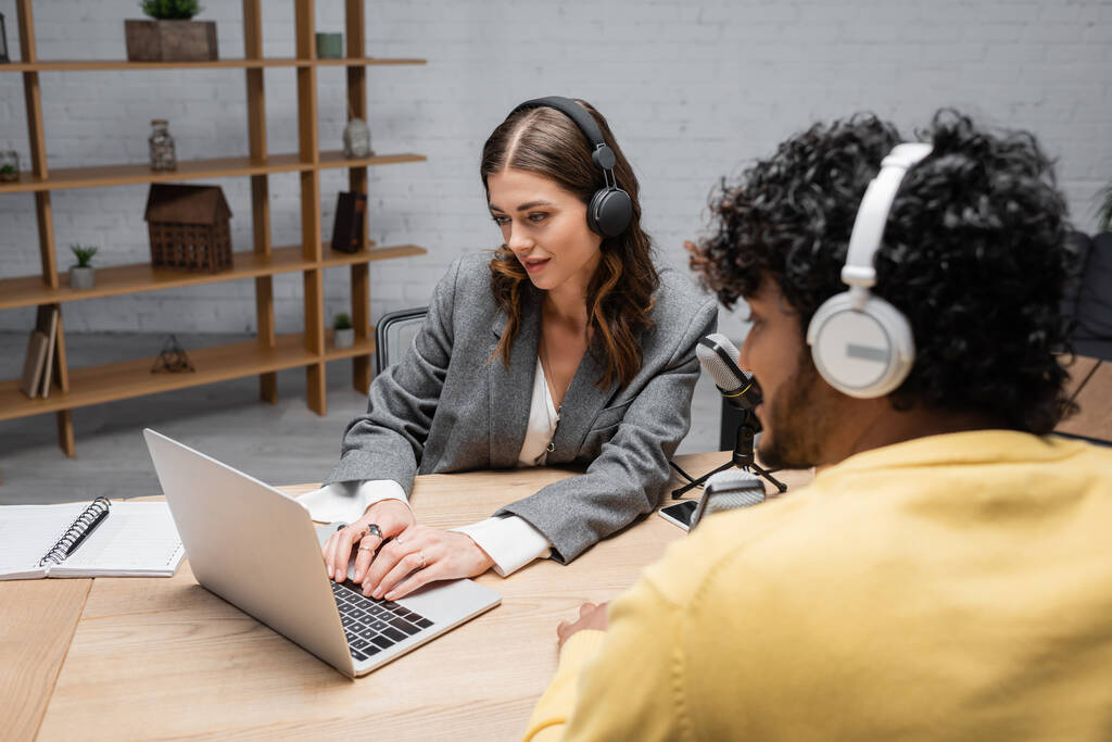 glimlachende brunette omroep in grijze blazer typen op laptop in de buurt van microfoons en notebook, terwijl wazig indiaanse man in gele jumper zitten in hoofdtelefoon in de radio studio - Foto, afbeelding