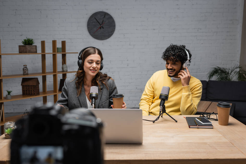 cheerful interracial podcasters in headphones working near professional microphones, notebook and mobile phone with blank screen on table with laptop and paper cups in modern studio - Photo, Image