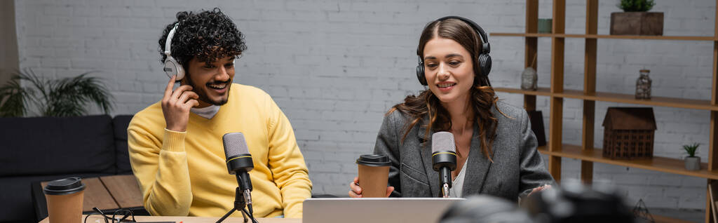 wesoły międzyrasowych nadawców w słuchawkach, żółty sweter i szary blezer patrząc na laptopa w pobliżu mikrofonów i kawy, aby przejść podczas pracy w nowoczesnym studio radiowym, baner - Zdjęcie, obraz