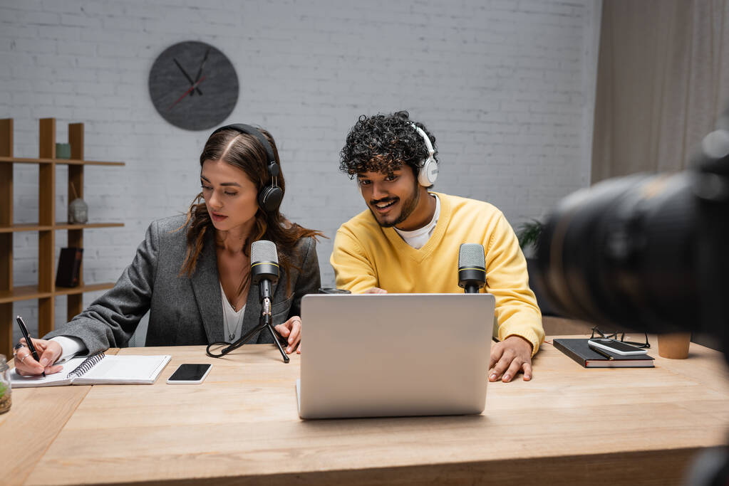 brunette radio host in headphones writing in notebook close to positive indian colleague looking at laptop near professional microphones and smartphone with blank screen in studio - Photo, Image