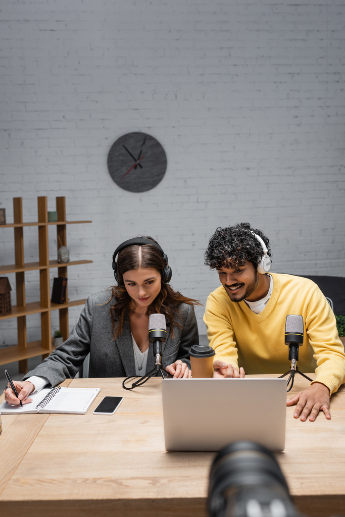 mosolygós podcaster fejhallgató és blézer írás notebook és nézi laptop közelében indiai kolléga sárga jumper, mikrofonok, kávé menni, és okostelefon a műsorszóró stúdióban - Fotó, kép
