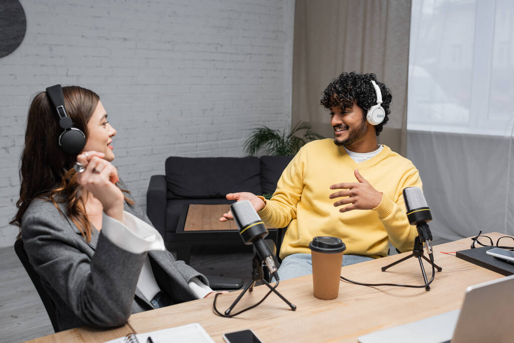 boldog multietnikus rádió házigazdák fülhallgató közelében beszélő professzionális mikrofonok, kávé menni, notebook és okostelefonok az asztalon a modern műsorszóró stúdióban karosszék a háttérben - Fotó, kép