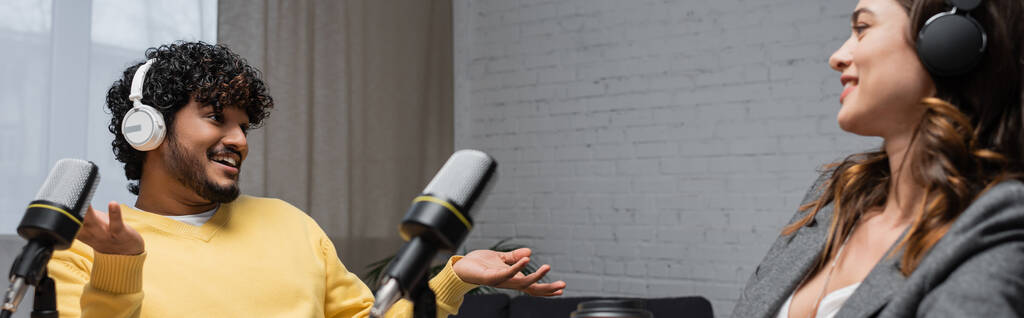 uomo indiano riccio e allegro in cuffia e maglione giallo parlando e gesticolando vicino a un collega bruna sorridente e microfoni professionali in studio radio, banner - Foto, immagini