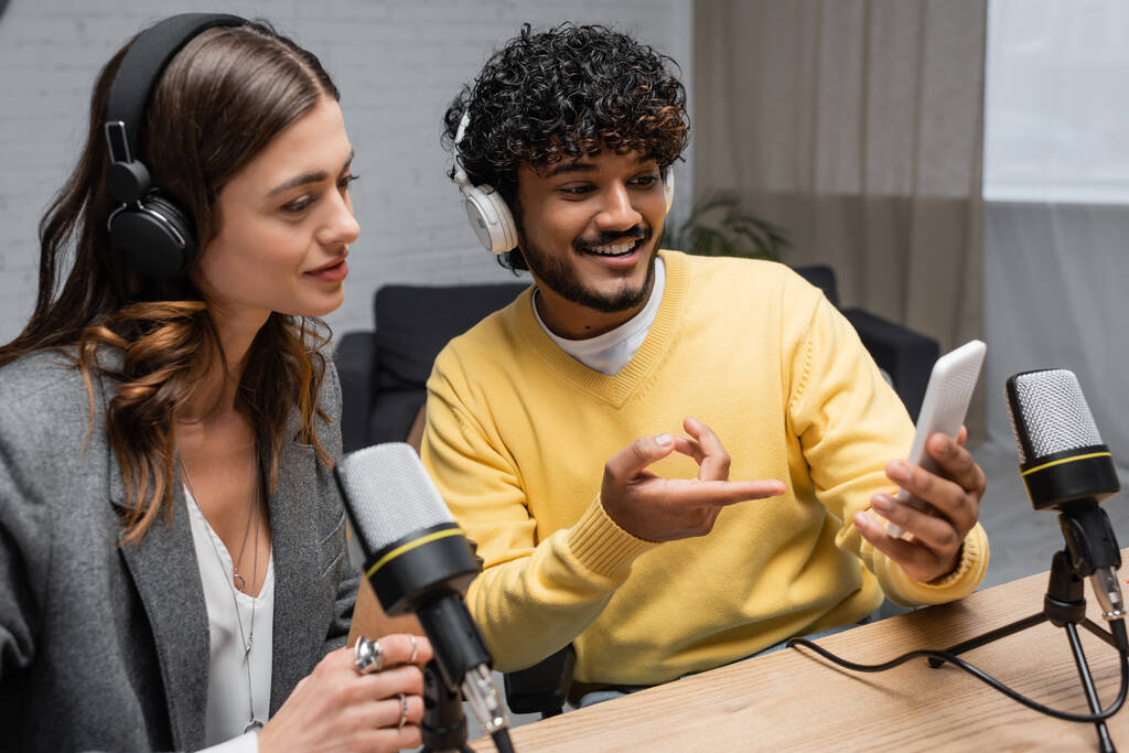 glimlachende indiase podcaster in koptelefoon en gele jumper tonen mobiele telefoon aan charmante brunette collega in de buurt van professionele microfoons in omroepstudio - Foto, afbeelding
