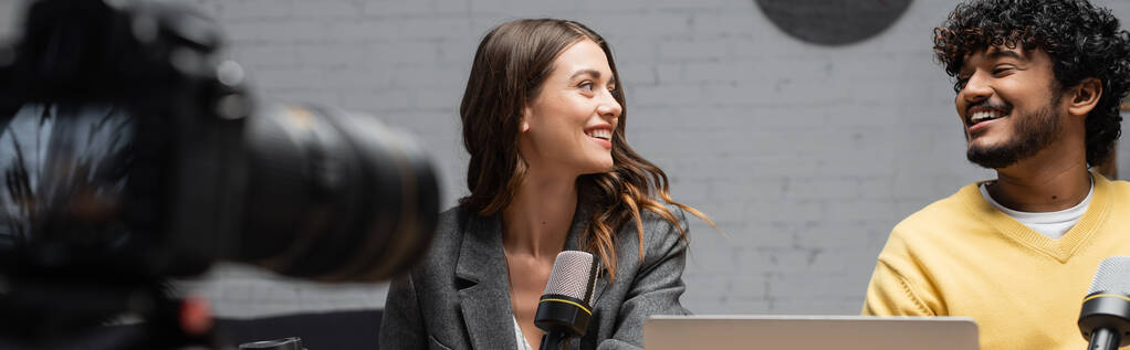 vousatý indián muž ve žlutém svetru a okouzlující brunetka žena v šedém blejzru usmívat se na sebe při nahrávání podcast před studio mikrofony a rozmazané digitální fotoaparát, banner - Fotografie, Obrázek