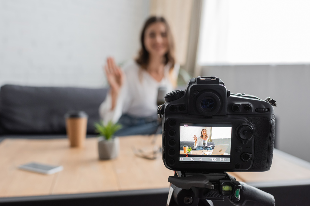 soustředěný snímek profesionálního digitálního fotoaparátu před rozmazanou ženou mávající rukou při nahrávání podcastu u papírového kelímku, smartphonu a květináče na stole ve studiu - Fotografie, Obrázek