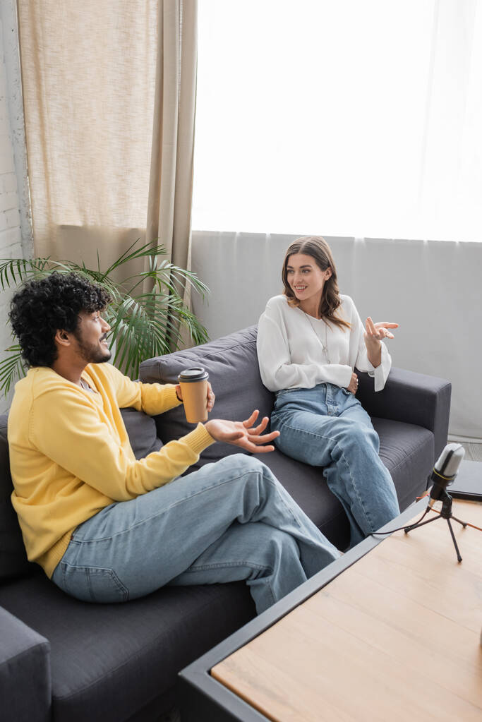 Hochwinkelaufnahme eines fröhlichen indischen Mannes mit Pappbecher und Gespräch mit einer Frau in weißer Bluse, während er auf einem Sofa in der Nähe von Notizbuch und Mikrofon im Podcast-Studio sitzt - Foto, Bild