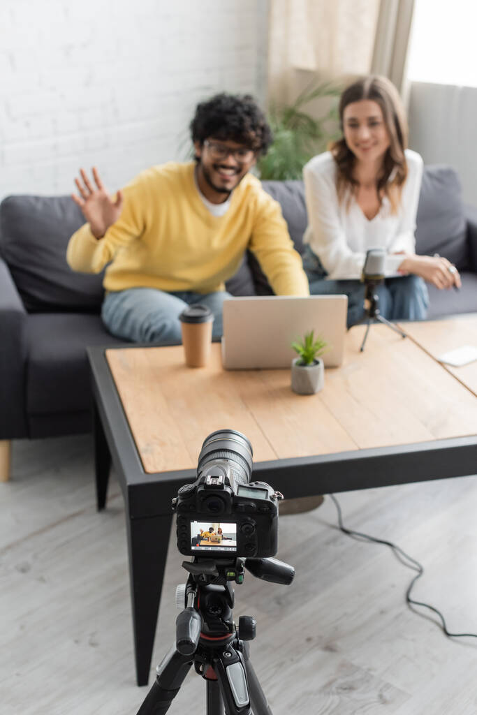 розмитий індійський чоловік махає рукою під час відео чату на ноутбуці біля усміхненого колеги і професійної цифрової камери в радіостудії з паперовою чашкою і вазоном на столі
 - Фото, зображення