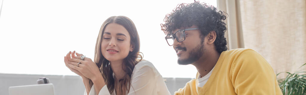 charming brunette woman and curly indian man in eyeglasses and yellow jumper looking at blurred laptop and smiling during video call in podcast studio, banner - Photo, Image