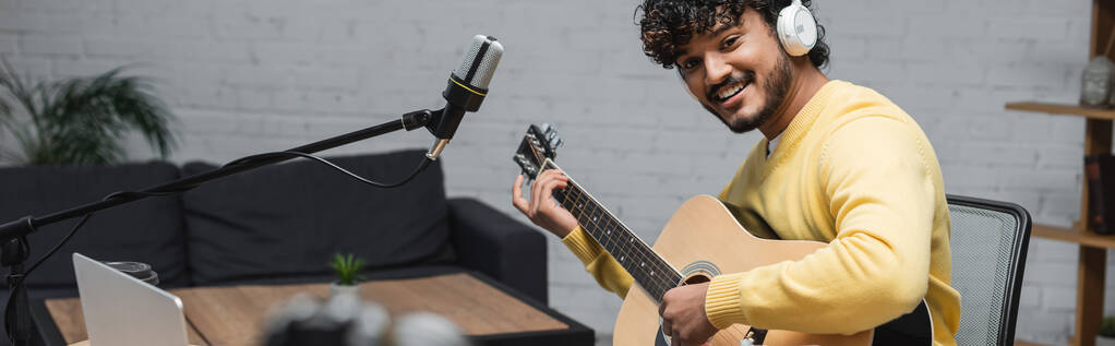 kręcone i szczęśliwy indyjski muzyk w słuchawkach i żółty skoczek gra na gitarze akustycznej w pobliżu laptopa i profesjonalny mikrofon w studio z sofą na tle, baner - Zdjęcie, obraz