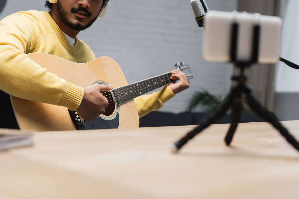 vista recortada del músico indio creativo en puente amarillo tocando la guitarra acústica cerca del micrófono y el teléfono inteligente con trípode mientras graba podcast en primer plano borroso - Foto, imagen