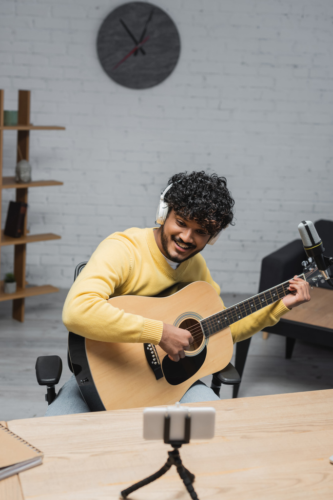 fröhlicher indischer Musiker mit Kopfhörer und gelbem Pullover spielt Akustikgitarre in der Nähe von verschwommenem Handy auf Stativ und professionellem Mikrofon, während er Podcast im Studio aufnimmt - Foto, Bild