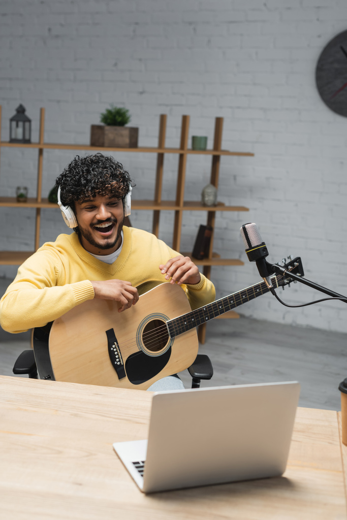 bouclé homme indien en casque sans fil et pull jaune rire tout en étant assis avec guitare acoustique près d'un ordinateur portable et microphones professionnels en studio - Photo, image