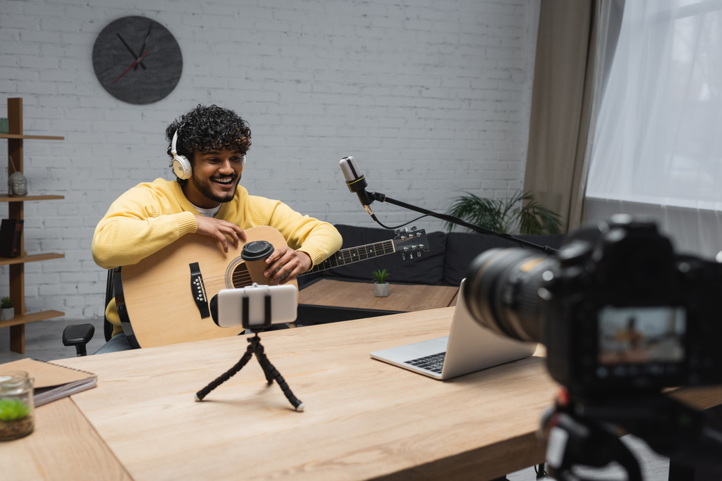 Podcaster indiano sorridente em fones de ouvido segurando guitarra acústica e café para ir perto de microfone e dispositivos durante o fluxo em estúdio de podcast  - Foto, Imagem