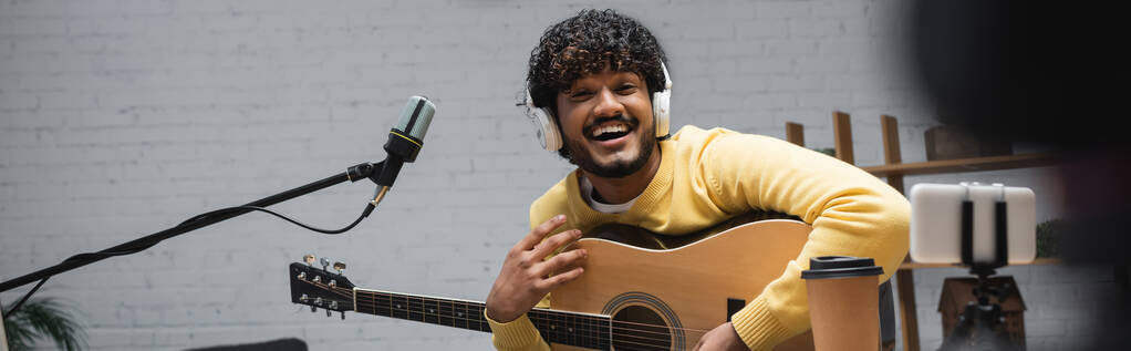 Веселый индийский подкастер в наушниках, держащий акустическую гитару рядом со смартфоном на штативе, кофе и микрофоне во время потока в студии, баннер  - Фото, изображение