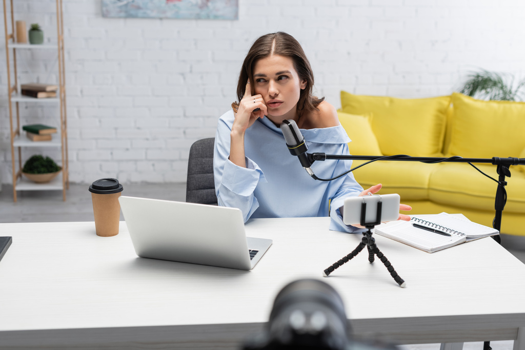 Pensive brunetka nadawca odwraca wzrok podczas rozmowy w pobliżu mikrofonu, smartfon na statywie, laptop i kawa, aby przejść do stołu podczas strumienia w studio podcast  - Zdjęcie, obraz