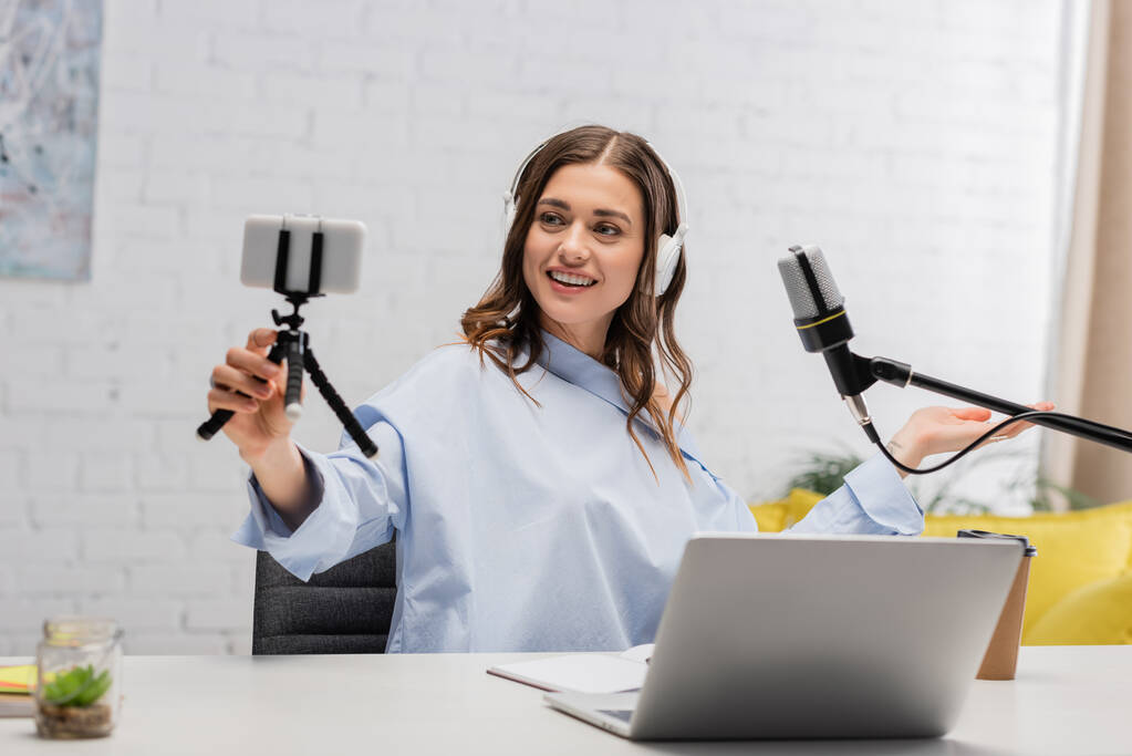 Brunetka podcaster w słuchawkach bezprzewodowych uśmiechając się podczas strumienia i trzymając smartfona na statywie w pobliżu mikrofonu i laptopa w studio  - Zdjęcie, obraz