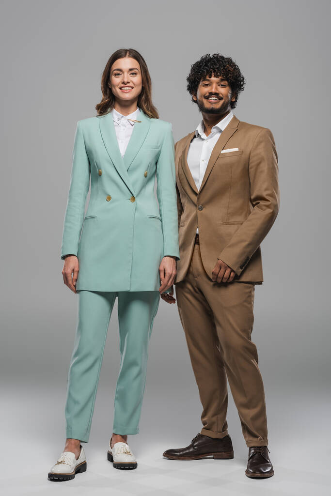 Full length of smiling multiethnic event hosts colleagues in formal wear looking at camera while standing together on grey background  - Photo, Image