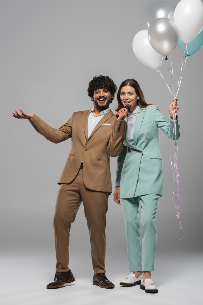 Полная длина позитивного индийского мероприятия в формальной одежде с микрофоном рядом с брюнеткой коллега держит праздничные воздушные шары на сером фоне  - Фото, изображение