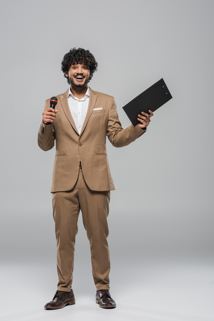 グレーの背景に立っている間、休日の間に無線マイクとクリップボードを保持茶色のスーツで笑顔インドのイベントホストの完全な長さ  - 写真・画像