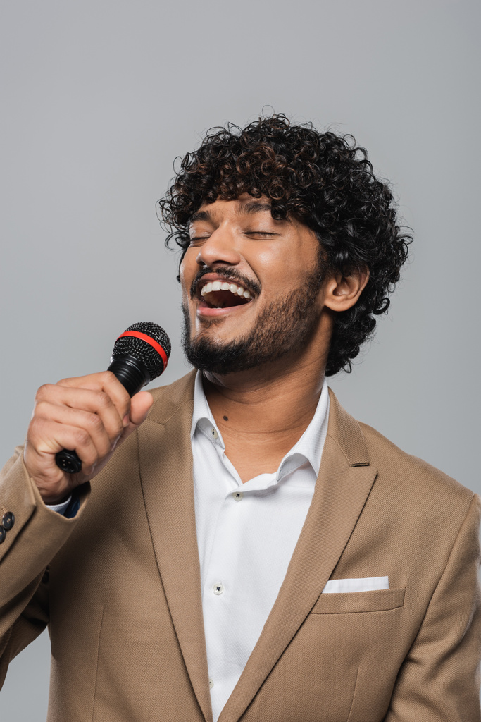 Portrait of cheerful indian event host with closed eyes in formal wear talking at microphone during performance while standing isolated on grey   - Photo, Image