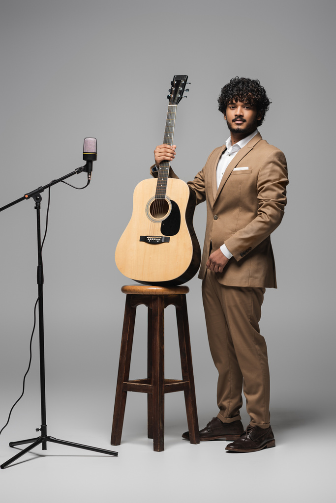 Повна довжина індійського ведучого заходу у формальному носінні, що тримає акустичну гітару біля стільця та мікрофона, дивлячись на камеру та стоячи на сірому фоні
  - Фото, зображення