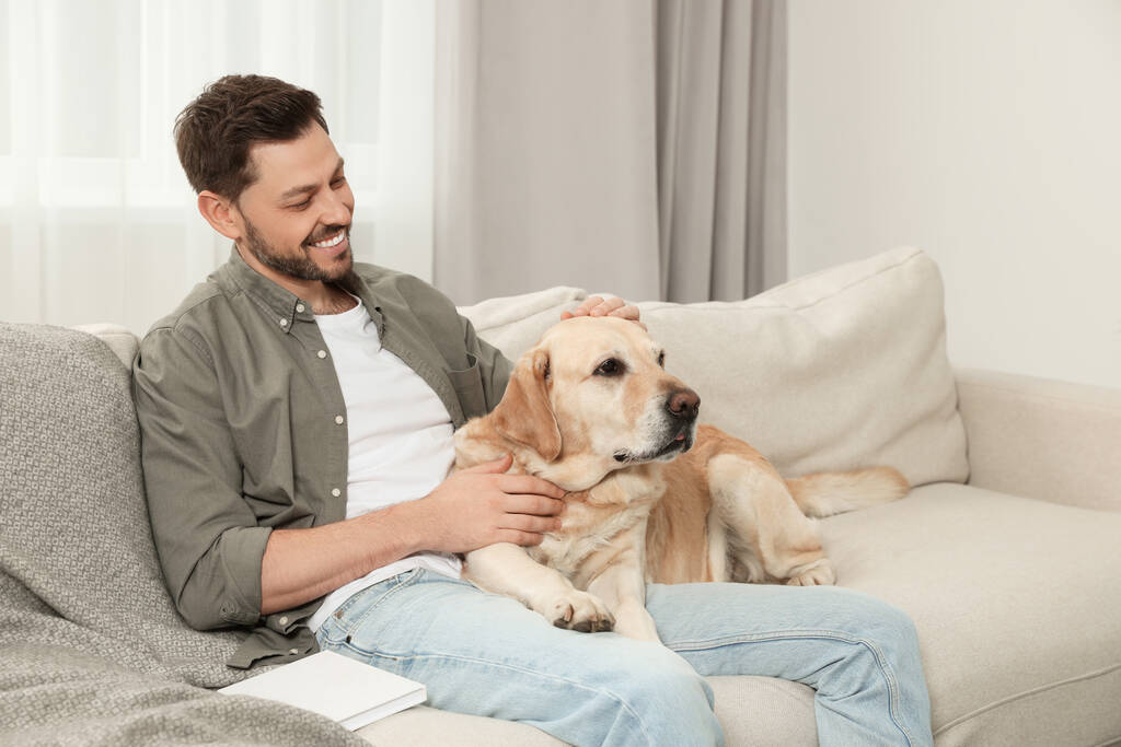 Hombre feliz sentado en el sofá con su lindo Labrador Retriever en casa - Foto, Imagen