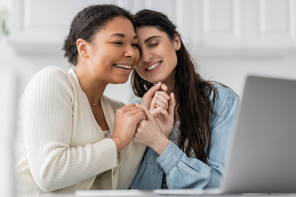 blij interraciale lesbisch paar holding handen in de buurt laptop  - Foto, afbeelding