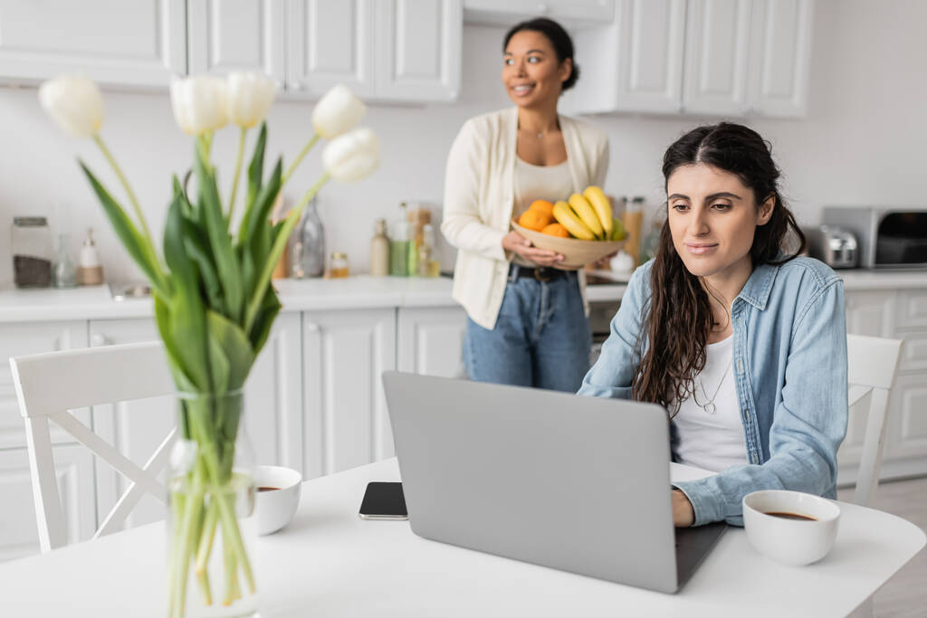 cheerful lesbian woman using laptop near multiracial girlfriend with bowl of fruits on blurred background  - Photo, Image