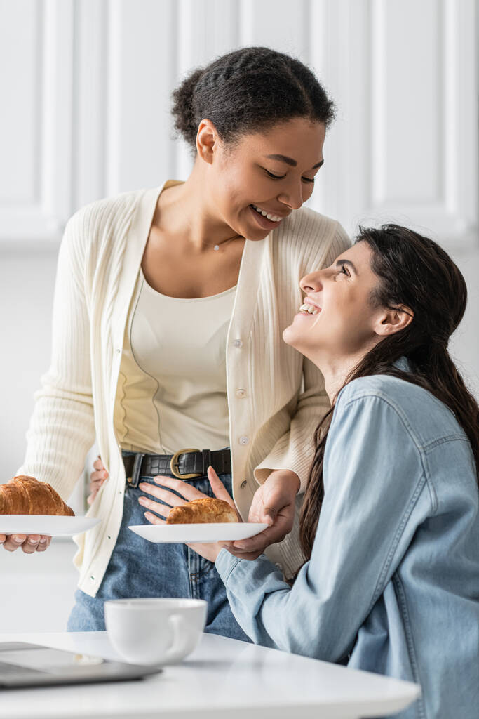 fröhliche und lesbische multirassische Frau, die Teller mit Croissants hält, während sie ihre glückliche Freundin ansieht  - Foto, Bild