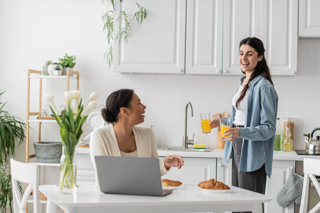 happy lesbian woman holding glasses with orange juice near multiracial girlfriend working on laptop  - Photo, Image