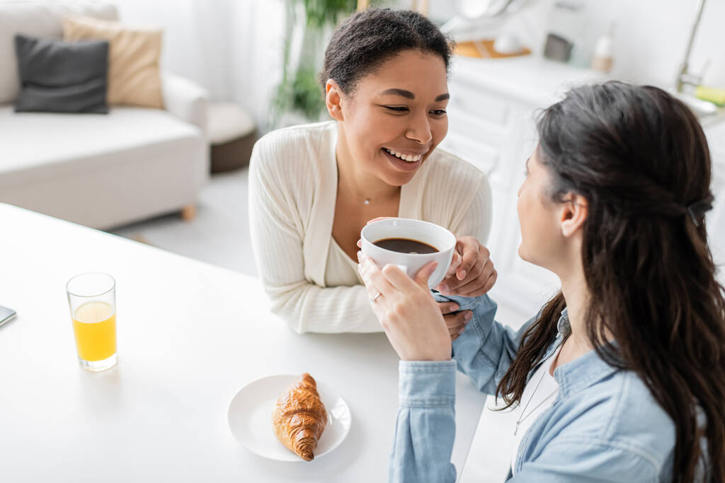 overhead view of cheerful multiracial lesbian couple smiling during breakfast in kitchen  - Photo, Image