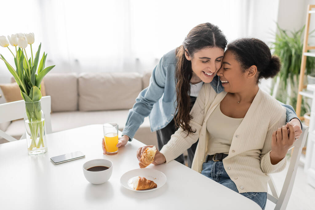 heureuse lesbienne femme tenant verre de jus d'orange et étreignant petite amie multiraciale pendant le petit déjeuner dans la cuisine  - Photo, image