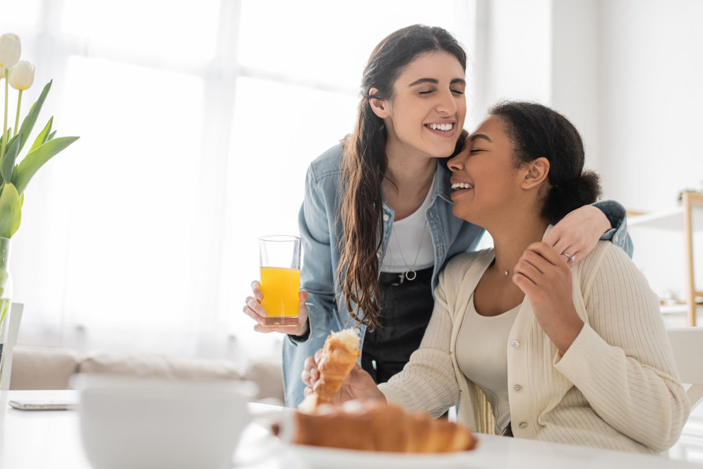 明るいレズビアン女性保持ガラスのオレンジジュースと巨大多人種のガールフレンドで朝食中にキッチン  - 写真・画像