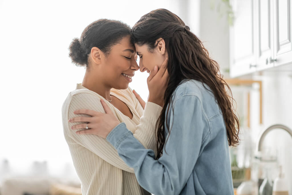 curly multiracial woman hugging cheerful girlfriend with closed eyes at home  - Photo, Image