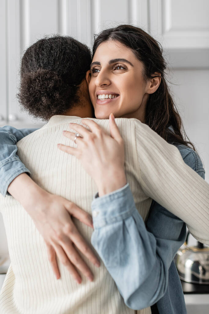 Parmağında alyansı olan mutlu lezbiyen kadın gülümsüyor ve çok ırklı kız arkadaşına sarılıyor.   - Fotoğraf, Görsel
