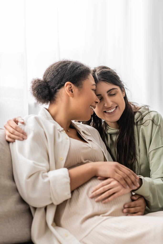 vrolijk lesbisch vrouw knuffelen zwanger multiraciale vrouw terwijl zitten op bank  - Foto, afbeelding