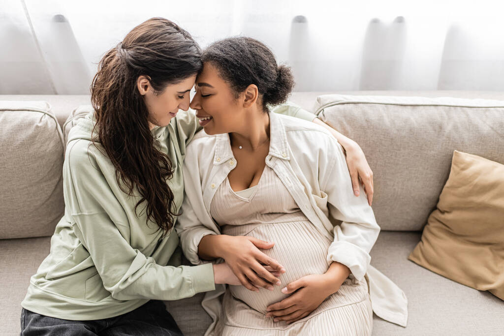 heureuse lesbienne femme câlin enceinte multiracial femme tandis que assis sur canapé  - Photo, image