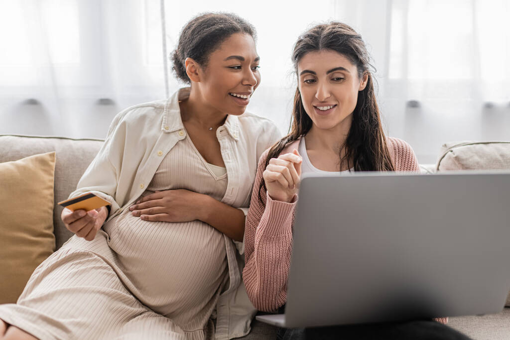 embarazada mujer multirracial celebración de la tarjeta de crédito mientras que hace compras en línea con alegre pareja lesbiana  - Foto, Imagen