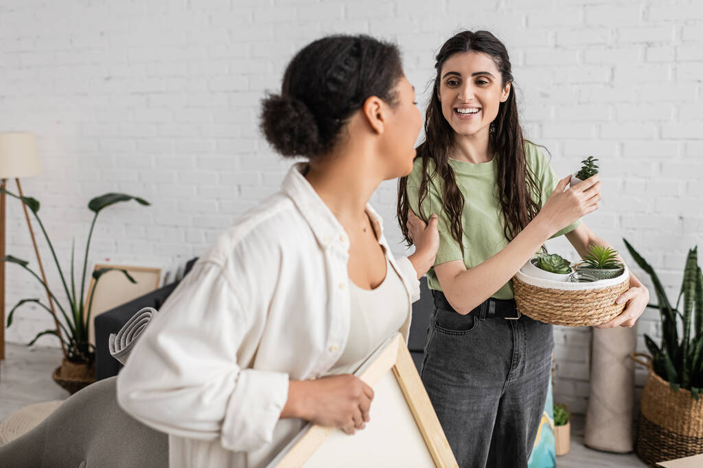 cheerful lesbian woman holding wicker basket with plants while looking at multiracial girlfriend during relocation to new house  - Photo, Image
