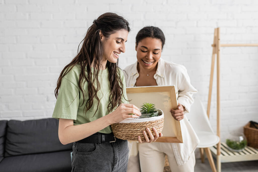 fröhliche lesbische Frau hält Weidenkorb mit Pflanzen neben glücklicher multirassischer Freundin in neuem Haus  - Foto, Bild
