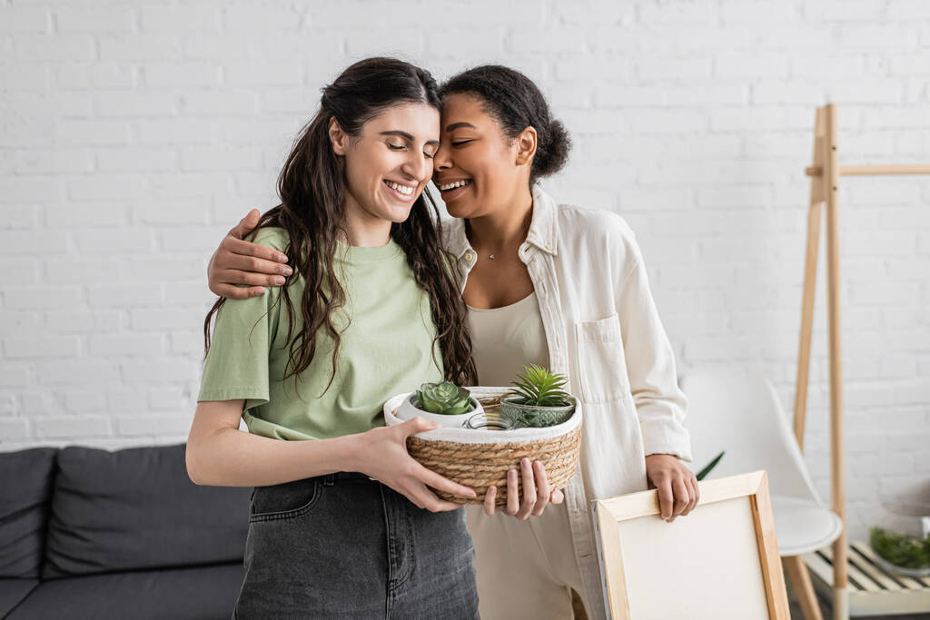 happy multiracial woman hugging cheerful lesbian girlfriend holding wicker basket with plants in new house  - Photo, Image