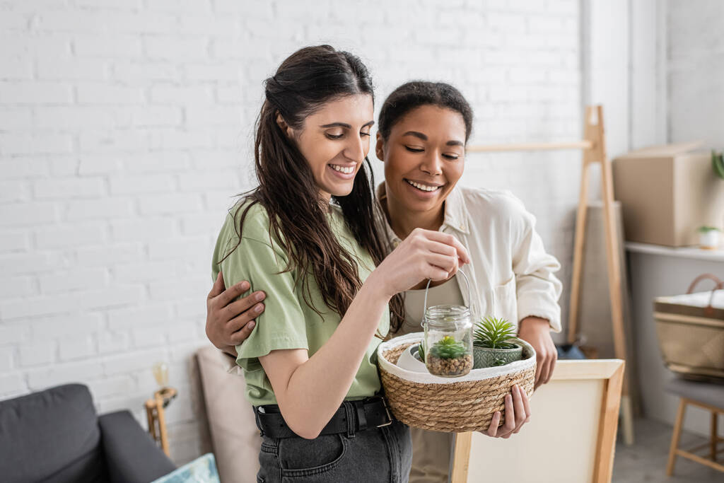 alegre lesbiana mujer celebración de mimbre cesta y vidrio tarro con plantas junto a positivo multiracial novia en nueva casa  - Foto, imagen