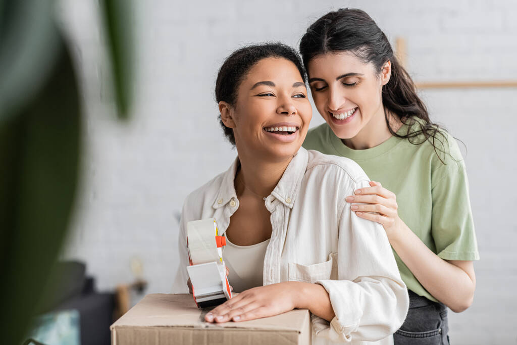 alegre multirracial mujer celebración cinta dispensador cerca caja de cartón y riendo con lesbiana pareja  - Foto, imagen