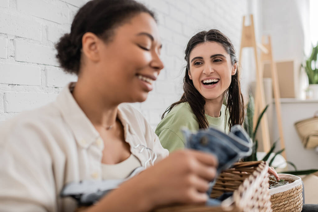 joyful lesbian woman looking at multiracial girlfriend folding clothes on blurred foreground  - Photo, Image