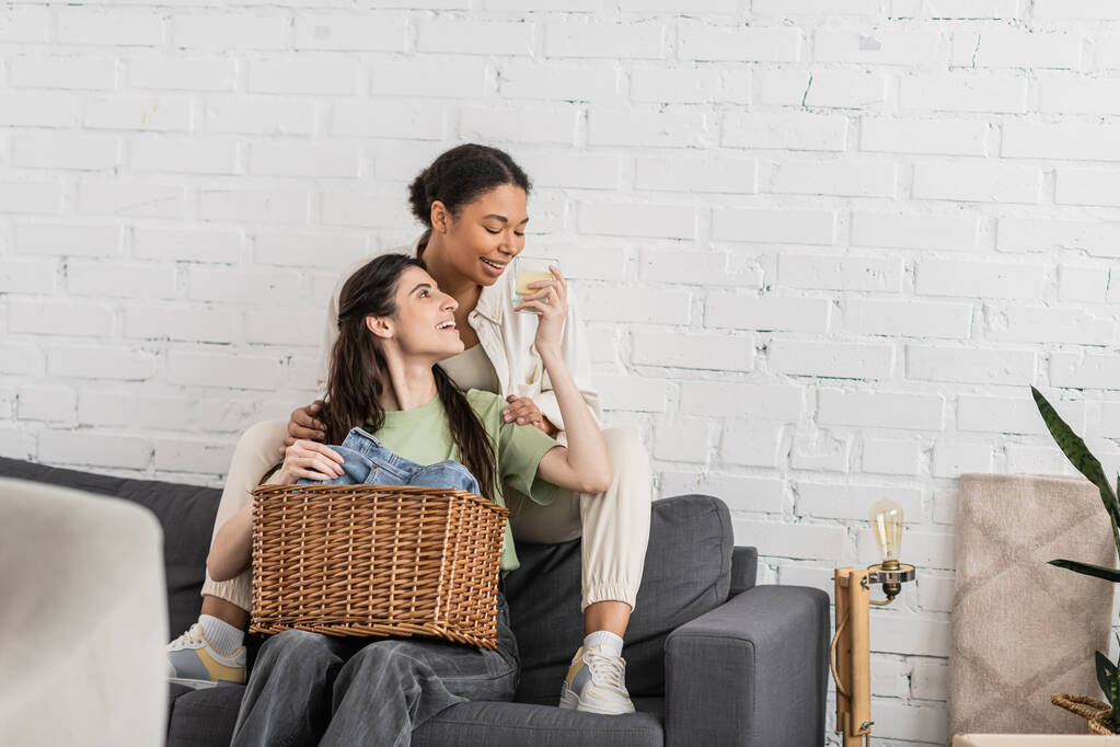 joyful lesbian woman holding scented candle while sitting on sofa next to multiracial girlfriend   - Photo, Image