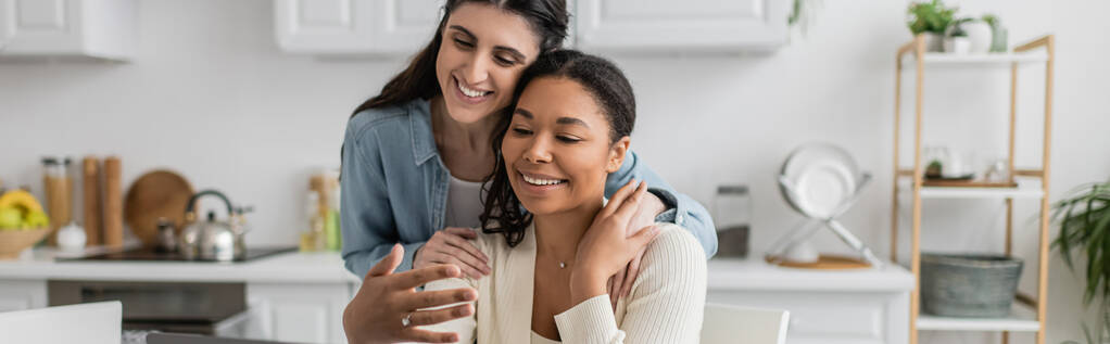 glücklich interracial lesbisches Paar umarmt einander in Küche, Banner - Foto, Bild