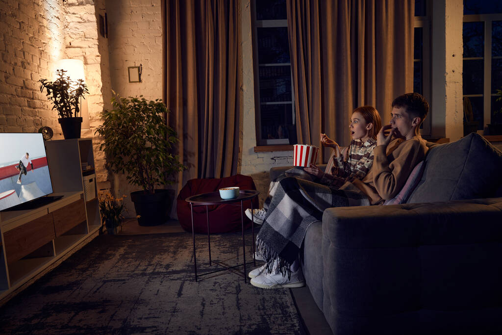 Jeune couple émotionnel, homme et femme passent du temps ensemble à la maison le soir, assis sur le canapé et regardant l'émission de télévision. Concept de relation, loisir, week-end, plaisir, émotions - Photo, image