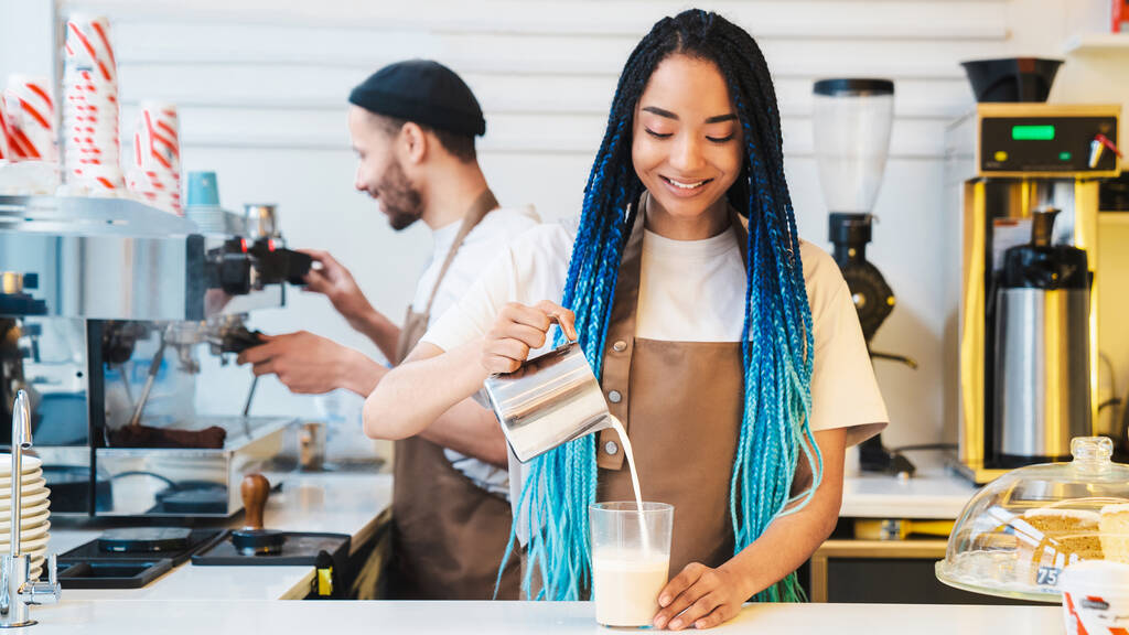 Barista femenina afroamericana haciendo una taza de café con leche mientras está detrás del mostrador de la cafetería. Dama vertiendo leche en el vaso - Foto, Imagen
