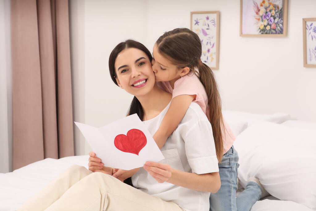 Mujer feliz con su hija y tarjeta de felicitación hecha a mano en la cama en casa. Celebración del día de la madre - Foto, imagen